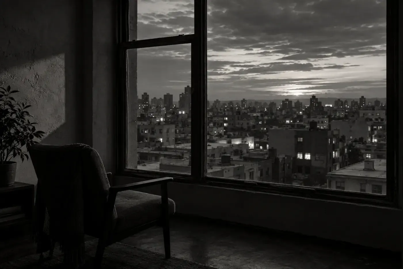 Fotografía en blanco y negro de una ciudad al atardecer vista desde una ventana, con un sillón en penumbra