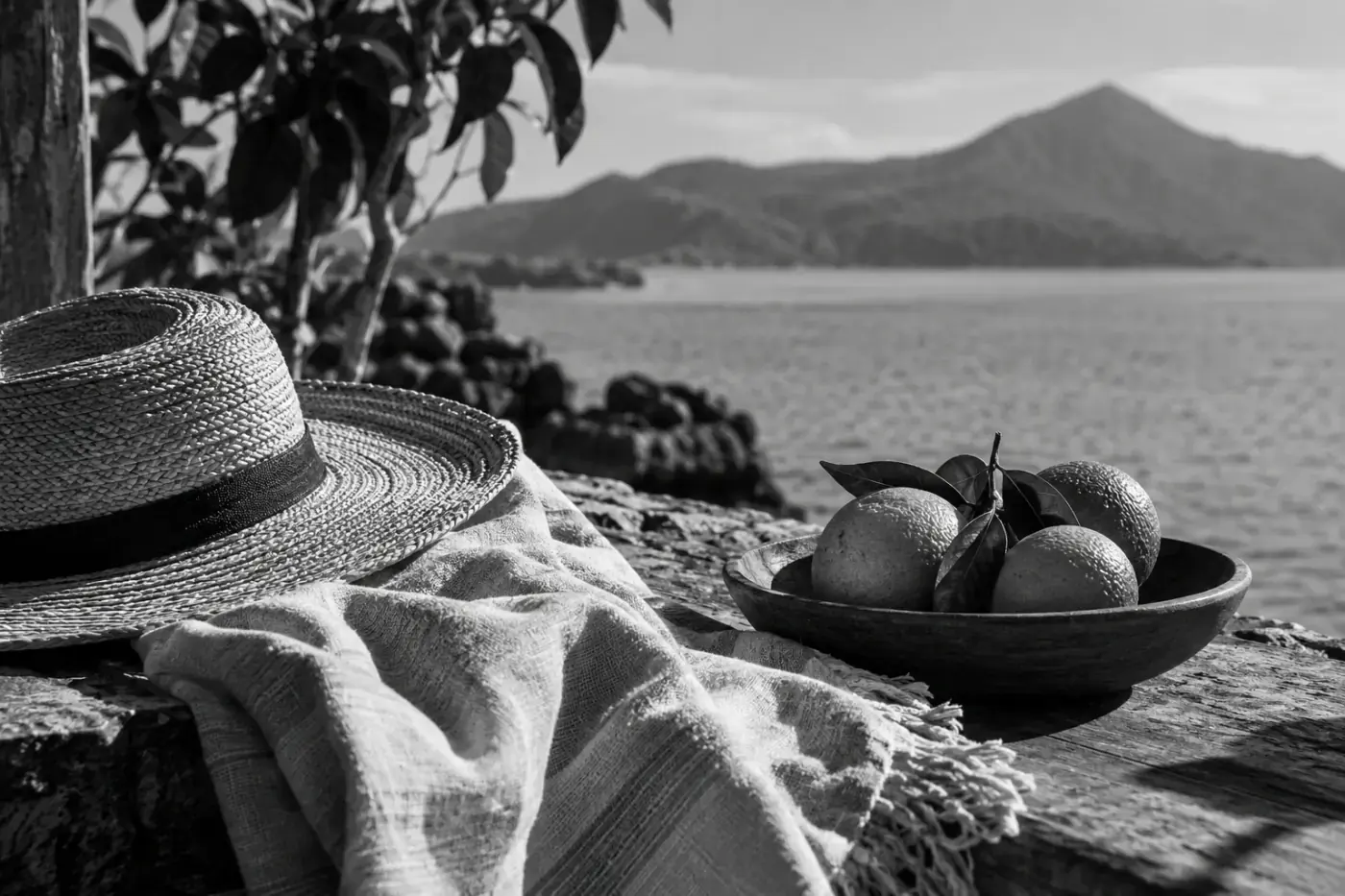 Fotografía en blanco y negro de un sombrero de paja, una manta y un cuenco con frutas frente a un paisaje de montañas y agua